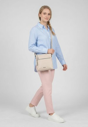 Junge Frau, die mit einer beigen Umhängetasche geht, trägt ein blau kariertes Hemd, pinke Hose und weiße Turnschuhe vor einem einfarbigen grauen Hintergrund.