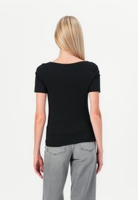 T-shirt nero aderente con maniche corte e scollatura ampia. Il tessuto appare liscio ed elastico. Il modello indossa jeans grigi.