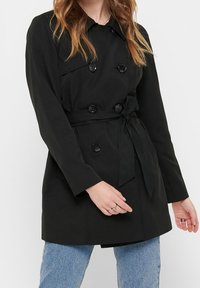 Schwarzer Trenchcoat mit doppelt geknöpftem Design, Fronttaschen und gebundenem Taillenbereich. Aus einem glatten Stoff gefertigt und mit großen Knöpfen ausgestattet.