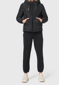 Sort pufferjakke med hætte, der har lynlåslukning foran, to sidelommer og elastiske manchetter. Sammen med sorte sweats og lyse sneakers.