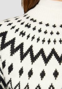 Witte gebreide trui met zwarte zigzag- en diamanten patronen, voorzien van een hoge kraag en geribbelde textuur, met contrasterende kleuren.