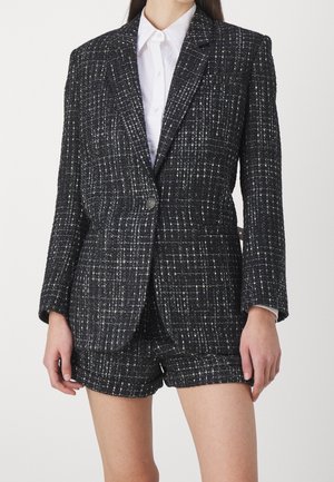 Vrouw draagt een zwart-wit gestructureerde geruite blazer met bijpassende korte broek en een wit overhemd met kraag eronder.