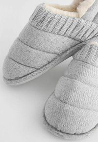 Graue Hausschuhe mit einem gesteppten Textildesign, weicher Innenausstattung und gerippten Strickakzenten am Kragen. Flache Gummisohle.
