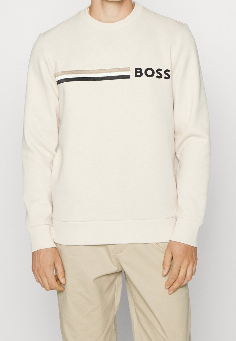 Homme portant un sweat-shirt crème avec des rayures beige, blanches et noires et le mot BOSS sur la poitrine, associé à un pantalon beige.