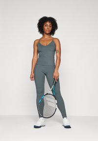 Szürke sportmellény és illő leggins ribbed szövetből. A modell teniszező ütőt tart, lábán fehér és kék sportcipő.