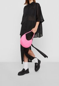 Růžová kabelka v podobě půlměsíce s hladkou texturou, kterou nese osoba v černé, volné košili a mokasínách s bílými ponožkami.