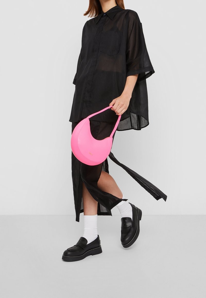 Rosa handväska i form av en månskära med slät yta, buren av en person som har på sig en svart, oversized skjorta och loafers med vita strumpor.