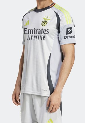 Biała sportowa koszulka z czarnymi i neonowo-żółtymi detalami, z logo Emirates Fly Better i Betano, noszona z dopasowanymi białymi szortami.