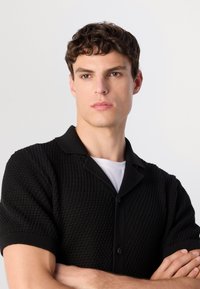 Jonge man met krullend bruin haar, gekleed in een zwarte gestructureerde kortebloes, over een witte T-shirt, met gekruiste armen en een neutrale uitdrukking.