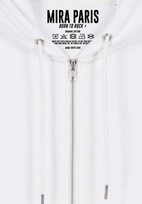 Felpa bianca con zip realizzata in cotone biologico, dotata di una zip argentata, lacci bianchi e un'etichetta stampata con le istruzioni per la cura.