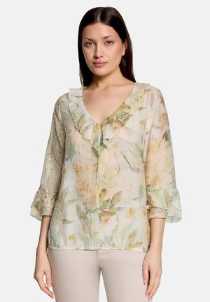 Femme portant un chemisier transparent à motif floral avec un col en V à volants et des manches trois-quarts, associé à un pantalon beige clair, debout devant un fond blanc.