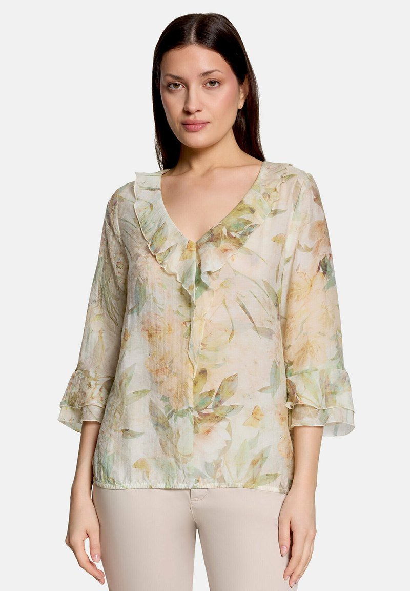 Femme portant un chemisier transparent à motif floral avec un col en V à volants et des manches trois-quarts, associé à un pantalon beige clair, debout devant un fond blanc.