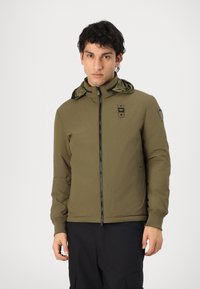 Veste vert olive avec fermeture éclair à l'avant, poignets élastiques et capuche à motifs. Dotée d'une fermeture éclair noire et de patchs sur les manches. Texture lisse.