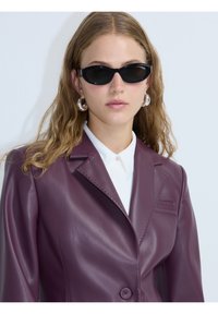 Blazer in pelle viola con rever a punta, chiusura a un bottone e dettagli cuciti. Occhiali da sole ovali neri e camicia bianca sotto.