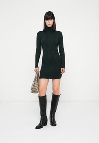 Vestido de tubo de color verde oscuro, hasta la rodilla, combinado con botas negras hasta la rodilla y un bolso de mano con estampado de piel de serpiente texturizado.
