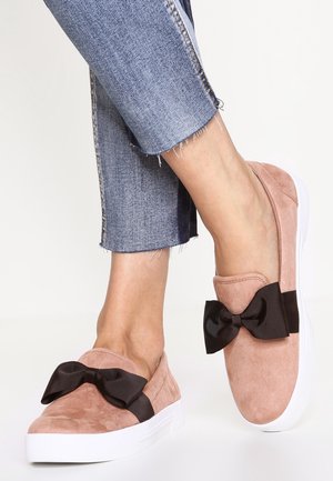 Pieds portant des chaussures en daim rose à enfiler avec des semelles blanches et de grands nœuds noirs, associées à un jean court en denim bleu.