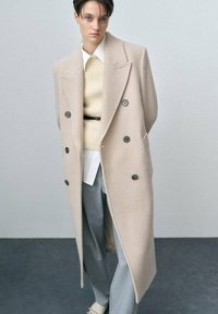 Cappotto in misto lana beige con design doppiopetto, ampie rever e bottoni scuri, indossato sopra un maglione chiaro e pantaloni grigi.