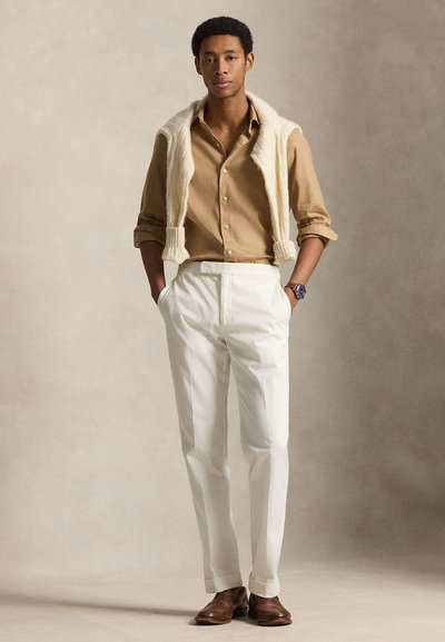 Άνδρας στέκεται με τα χέρια στις τσέπες, φοράει μπεζ πουκάμισο, λευκό παντελόνι, καφέ παπούτσια, κρεμ πουλόβερ περασμένο στους ώμους και ένα ρολόι χειρός.