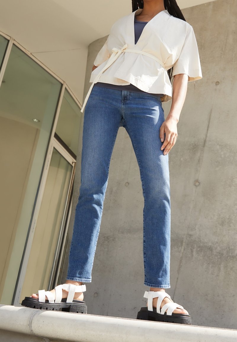 Haut crème en style peplum avec une taille à nouer, associé à un jean skinny bleu et des sandales noires à plateforme avec des brides blanches.