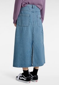 Falda de mezclilla azul clara de largo completo, con una abertura en la parte trasera y dos grandes bolsillos traseros. Combinada con zapatillas negras y blancas.