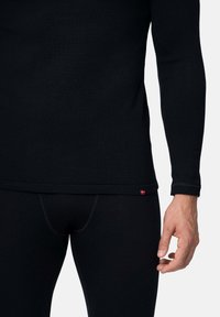 Top térmico negro de manga larga con un diseño ajustado y textura acanalada. Pequeña etiqueta de acento roja en el dobladillo. Combina con pantalones térmicos negros.