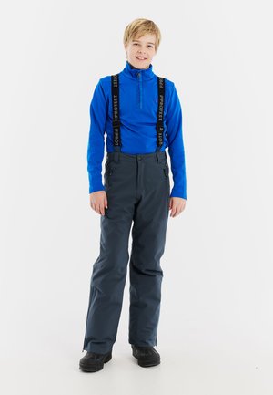 Blauwe long sleeve top met ritskraag, gecombineerd met donkere grijze waterdichte broek met verstelbare bretels en twee zijzakken. Zwarte laarzen.