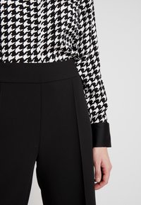 Sorte bukser med bred talje, parret med en sort og hvid houndstooth-mønstret bluse med lange ærmer og sorte manchetter.