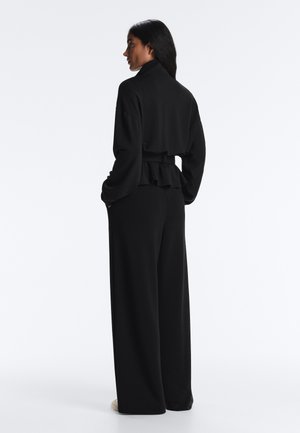 Pitkähiuksinen nainen seisoo mustissa leveälahkeisissa housuissa ja vastaavassa pitkähihkaisessa paidassa, jossa on vyöyksityiskohta, ja katsoo vasemmalle yksinkertaisen valkoisen taustan edessä.