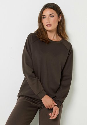 Donna con capelli castani alla lunghezza delle spalle che indossa una maglia a maniche lunghe marrone scuro e pantaloni coordinati, in piedi contro un muro bianco.