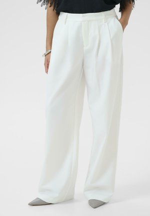 Ženska nosi bele široke hlače z visokim pasom, združene z sivimi čevlji s koničastimi konicami in črnim topom.