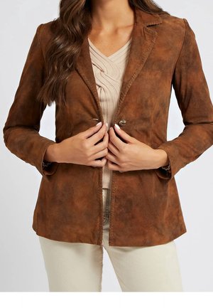 Kvinde iført en brun ruskindsblazer over en beige tekstureret top, der knapper blazerens enkeltknap, med cremefarvede bukser synlige.