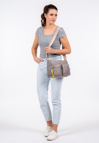 Borsa a spalla in tessuto grigio con due tasche frontali, tirazip giallo e tracolla beige regolabile, abbinata a jeans chiari e scarpe bianche.