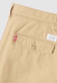 Pantalon en coton beige avec une poche arrière boutonnée, une étiquette cousue et une étiquette rouge. Tissu texturé avec des détails de surpiqûres subtils.