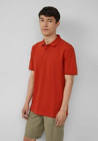 Rotes Polohemd mit Kragen, kurzen Ärmeln und dezentem Logo. Kombiniert mit hellgrünen Shorts, vor neutralem Hintergrund stehend.