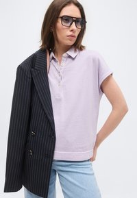 Γυναίκα που φοράει μεγάλα μαύρα γυαλιά ηλίου, ανοιχτό μωβ πόλο με κοντά μανίκια, ανοιχτό μπλε τζιν και κρατάει μια μαύρη σακάκι με ραβδώσεις στον ώμο.