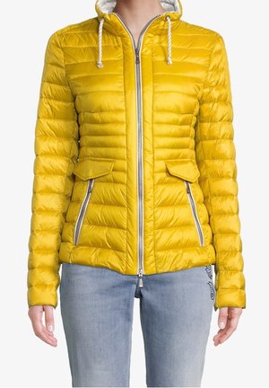 Kvinde iført en knaldgul, quiltet hættejakke med hvide snore og sølvfarvede lynlåse foran, kombineret med lyseblå jeans.