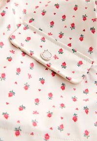 Tissu blanc avec un motif de petits boutons de rose roses, cœurs et papillons, avec un rabat de poche boutonné avec texte rouge sur le bouton.