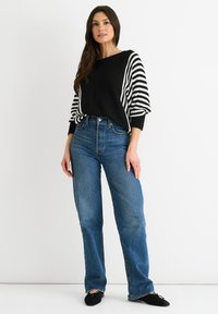 Maglione a righe bianco e nero con maniche ampie, abbinato a jeans blu a vita alta. Il modello è in piedi su uno sfondo neutro.