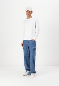 T-shirt blanc à manches longues avec un petit logo, associé à un pantalon cargo en denim bleu ample et des baskets blanches. La tenue est complétée par une casquette beige.