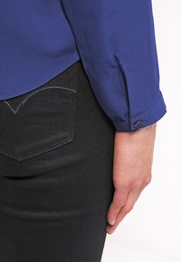 Blouse bleue à texture lisse avec des poignets boutonnés, associée à un jean noir ajusté présentant un design de poche diagonale et des coutures foncées.