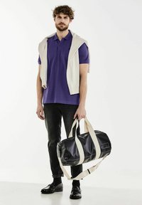 Sac de sport noir avec des poignées crème, doté d'une fermeture éclair. Le modèle porte un polo violet et un pull crème drapé sur les épaules avec un jean foncé.
