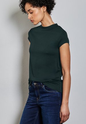 Dark green, short-sleeve t-shirt with a high neck, made of a smooth fabric. Worn with dark blue jeans, featuring a standard fit and slight stretch.