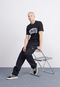 Černé tričko s potiskem "Dickies", černé cargo kalhoty a kostkované tenisky na slip-on. Sedí na chromově sklápěcím křesle proti jednobodovému pozadí.