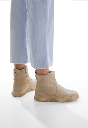 Bottines en suède beige avec un col en fourrure doublé, fermeture éclair sur le côté et semelle en caoutchouc texturé, présentées avec un pantalon cropped rayé.
