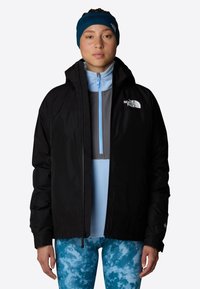 Czarna wodoodporna kurtka z zamkiem błyskawicznym, regulowanym kapturem i logo The North Face na lewej piersi. Nałożona na jasnoniebieską bluzkę.