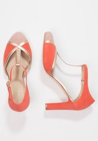 Korálově semišové vysoké podpatky se zlatým metalickým akcentem na špičce a pásku. Mají překřížené pásky a uzavřenou patu se zapínáním na kotníku.