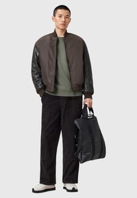 Chaqueta bomber marrón con mangas de cuero negro, sudadera verde oliva, pantalones de pana negros y una bolsa negra. Zapatos de color claro.