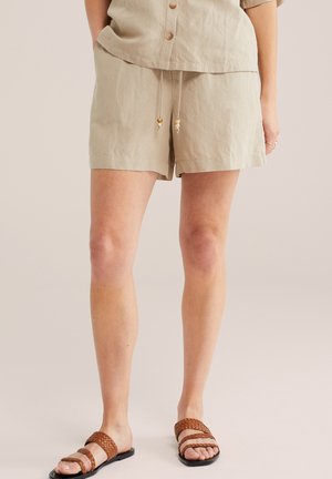 Vrouw draagt beige short met trekkoord en bijpassende korte mouw top, gecombineerd met bruine gevlochten platte sandalen op een neutrale achtergrond.