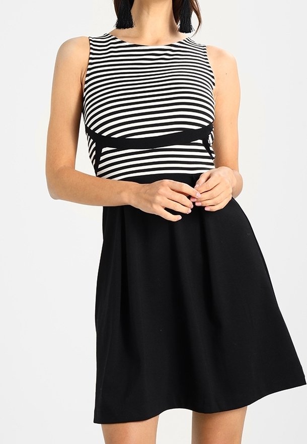 Femme portant une robe sans manches avec des rayures horizontales noires et blanches en haut et une jupe noire unie, mains jointes à la taille.
