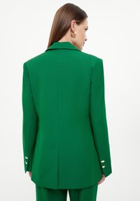 Blazer verde su misura con rever appuntiti, spalle strutturate e tre bottoni dorati su ciascun polsino. Tessuto dalla consistenza morbida.